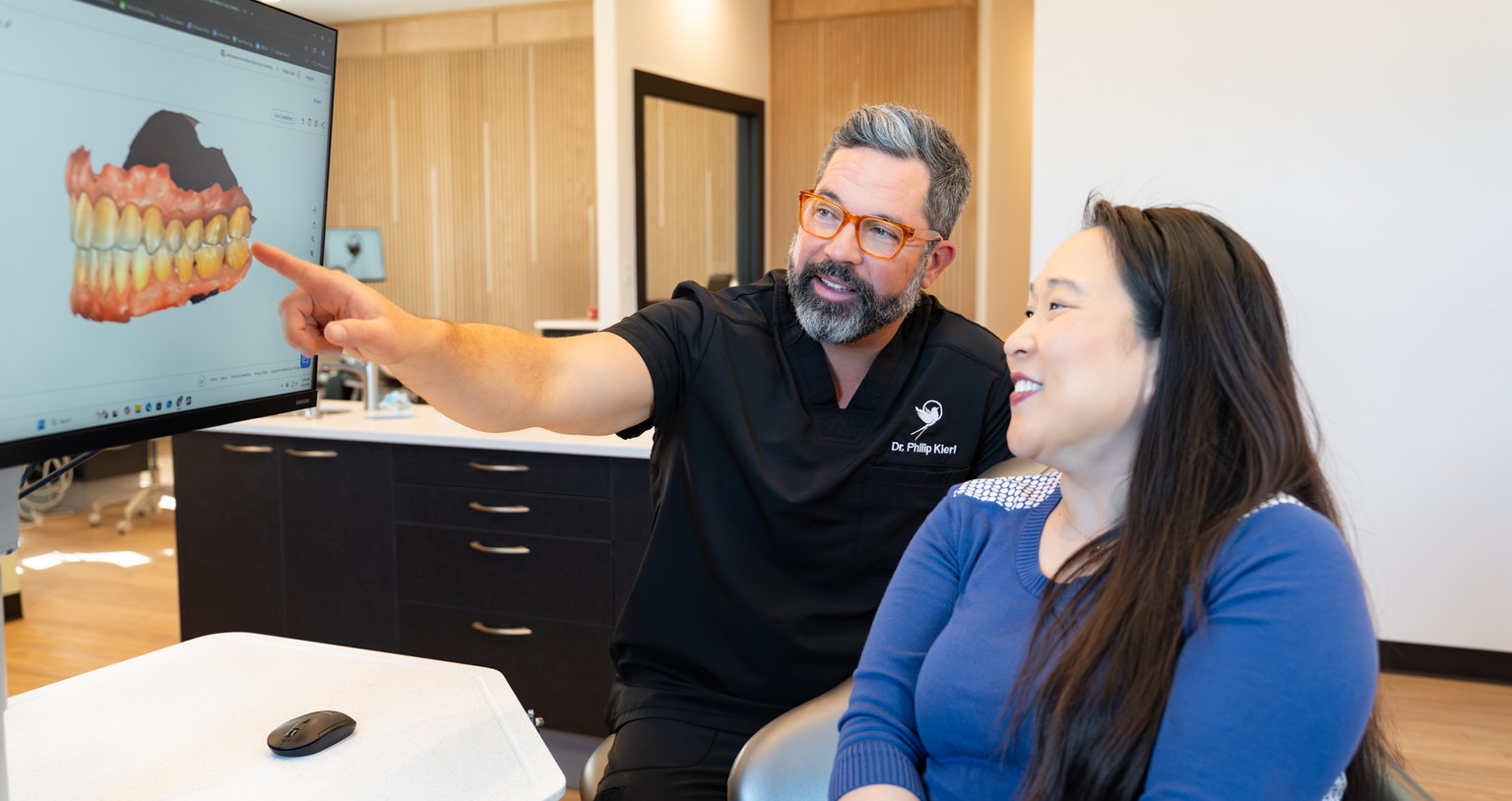 Scissortail orthodontist pointing at a 3D digital treatment plan on a screen while a smiling adult patient looks on.