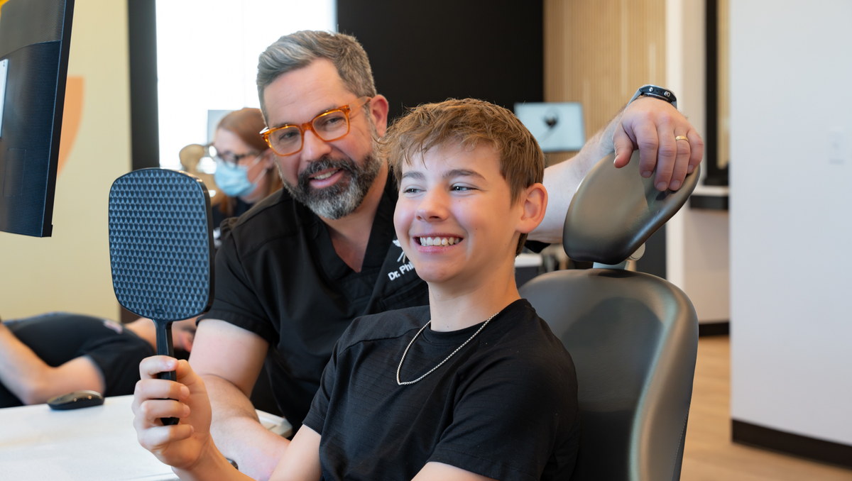 Teen patient smiling into a hand mirror while a Scissortail Orthodontics doctor stands beside the chair.
